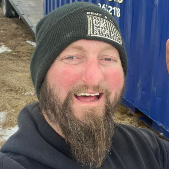 Close up head and shoulders photo of a man with a long beard, wearing a woolly hat with "CK Storage", in front of a blue storage container