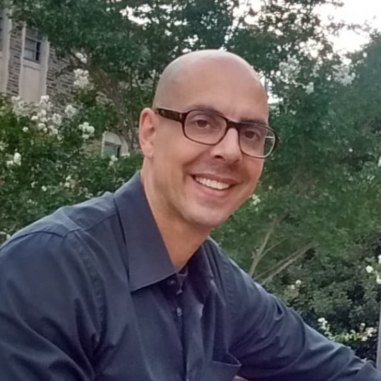 Head and shoulder portrait. Young man with shaved head and glasses, smiling at the camera. He wears a dark blue shirt. There are office buildings and trees in the background.
