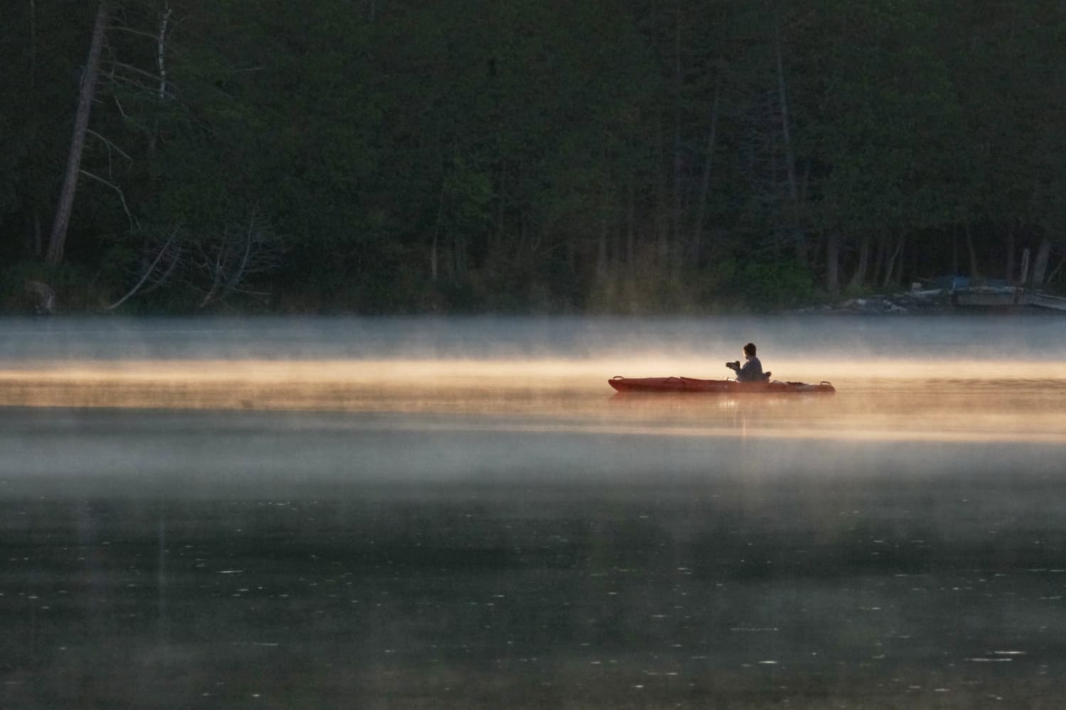 Telephoto image of a person in a red or brown kayak holding a camera, on a lake with early morning mist rising through a sunbeam. The water is dark in the foreground and the background show trees right down to the lake shore.