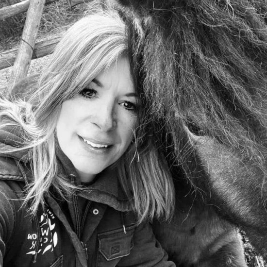 Head and shoulders black and white photo of a woman with long, fair hair, leaning in to the head of a pony with a dark coat. Some wooden fencing is visible behind them.