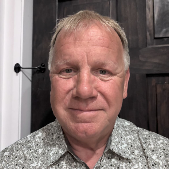 Head and shoulder photo of a man with greying hair, wearing a white and grey patterned shirt. He is standing in front of a dark wooden panelled door with a white frame.