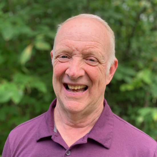 A head and shoulder photo of a man with grey hair wearing a bright purple open neck shirt, standing in front of out-of-focus green foliage.