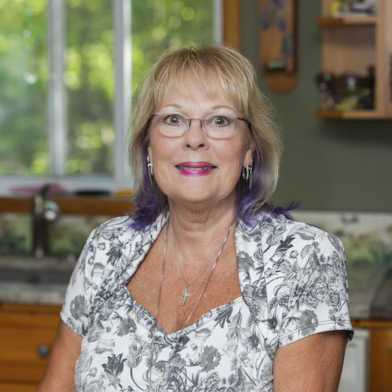 Woman with fair hair and purple tints is sitting in a litchen, in front of a window with trees outside. She wears glasses and a short-sleeved white top with a grey floral pattern.